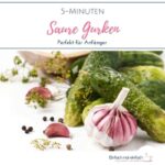 Saure Gurken mit Knoblauchknolle, Pfefferkörnern und Dillblüten neben einem hälzernen Messlöffel mit Salz.